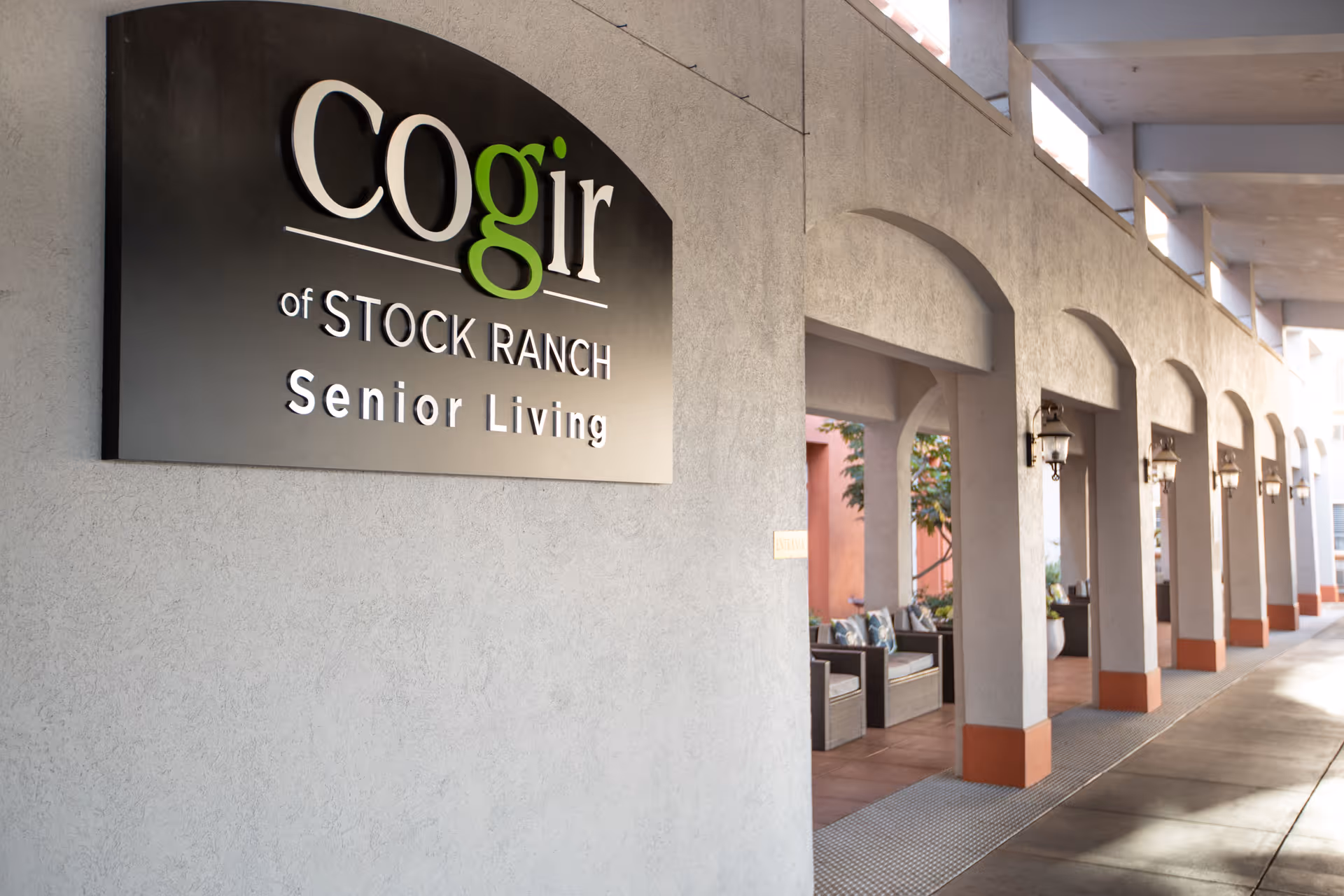 Exterior view of Cogir of Stock Ranch Senior Living facility showing a long covered walkway with arches, outdoor seating areas with chairs and cushions, and wall-mounted lantern-style lights.