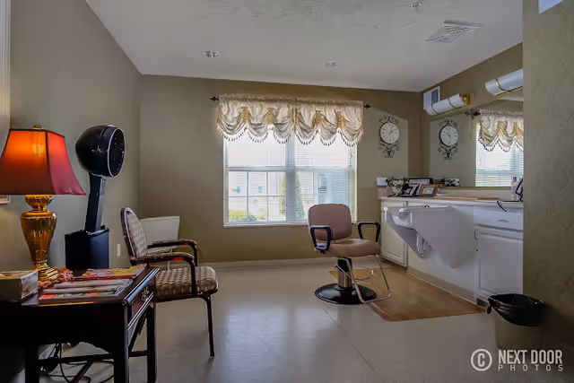 A well-lit hair salon room in a senior living facility with a styling chair in front of a large mirror and counter. There are two additional chairs, a table with magazines, a lamp, and a hair dryer. A window with decorative curtains lets in natural light.