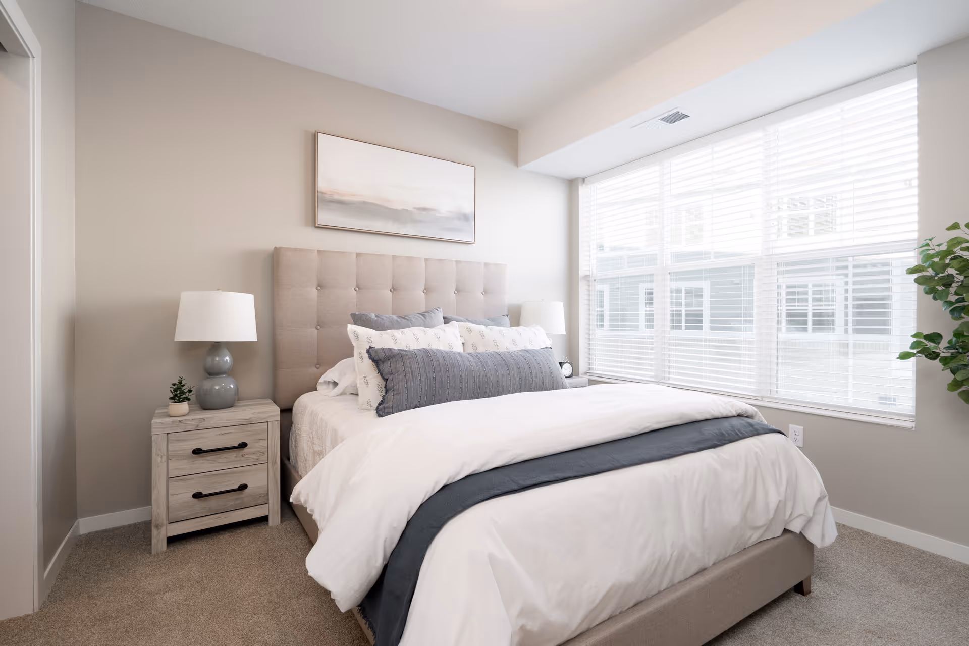 A bright and cozy bedroom with a large window covered by white blinds allowing natural light to fill the room. The bed has a tufted beige headboard and is made with white and gray bedding, including multiple pillows. On the left side of the bed is a wooden nightstand with two drawers, a gray lamp, and a small potted plant. A framed abstract painting hangs above the headboard, and there is a green leafy plant in the corner near the window.