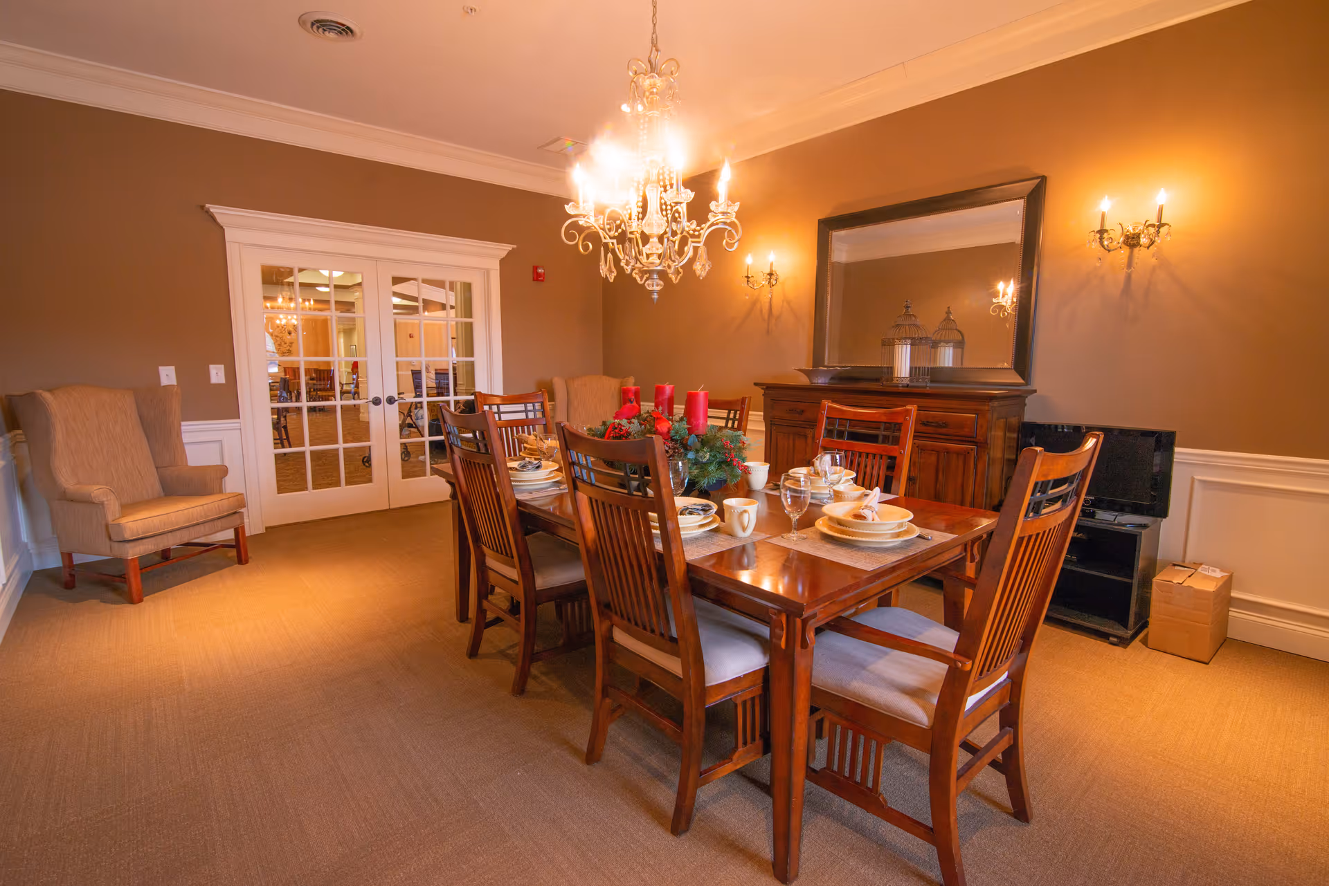 A warm dining room with a wooden table set for a meal, chandelier overhead, sideboard with mirror, and upholstered chairs.