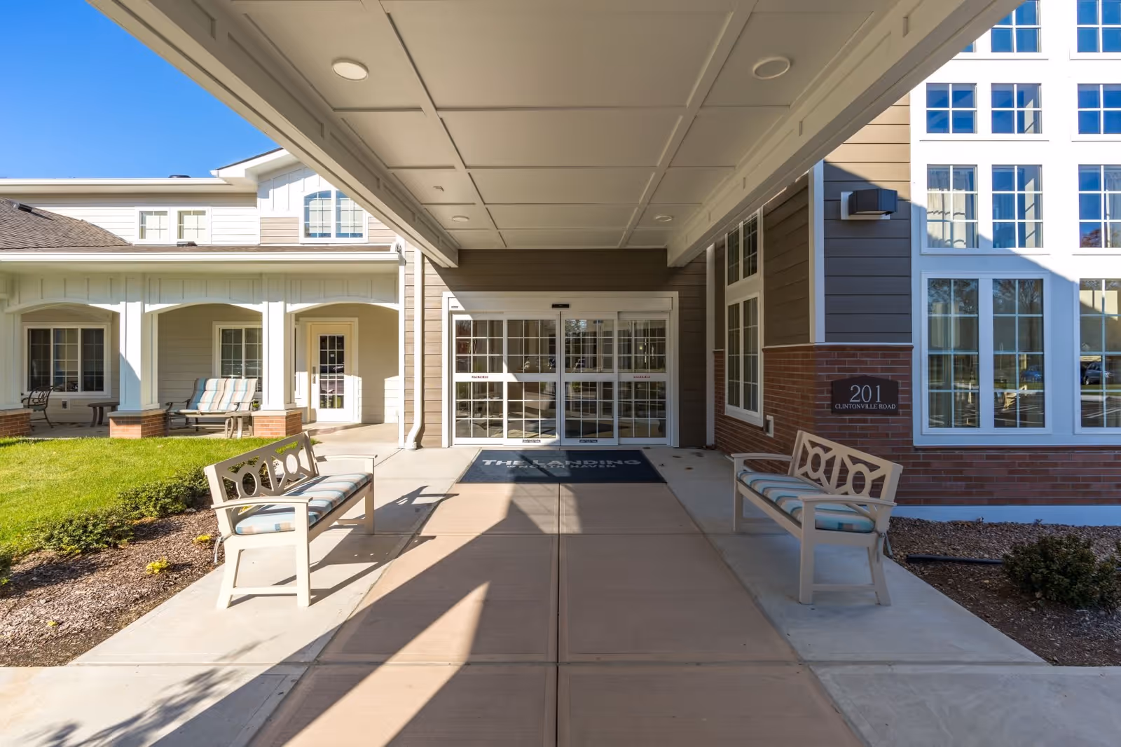 Entrance area of The Landing of North Haven facility with a covered walkway, two benches with cushions on either side, glass double doors, and a building sign displaying the address 201 Clintonville Road.