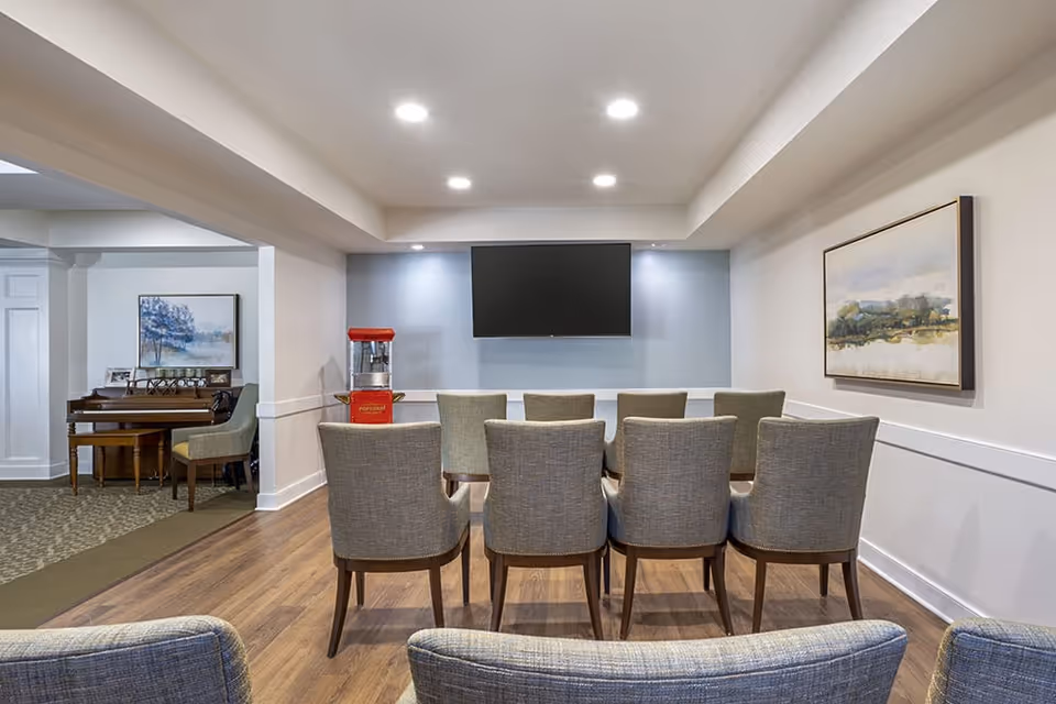 A small theater-style room with two rows of gray upholstered chairs facing a wall-mounted flat screen TV. To the left, there is a red popcorn machine and a piano with a chair. The room has wood flooring, white walls with a blue accent wall behind the TV, and framed artwork on the walls.