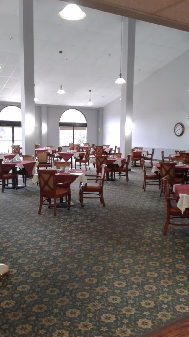 A spacious dining room with multiple wooden tables and chairs arranged neatly. Each table is covered with a white tablecloth and a burgundy overlay, set with glassware and napkins. The room has high ceilings with hanging pendant lights and large arched windows letting in natural light. A clock is mounted on the wall.