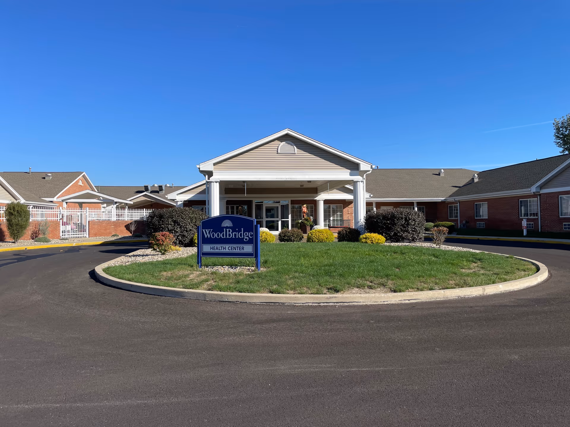 Front entrance of the WoodBridge Health Center with a circular driveway and landscaped island under a clear blue sky.