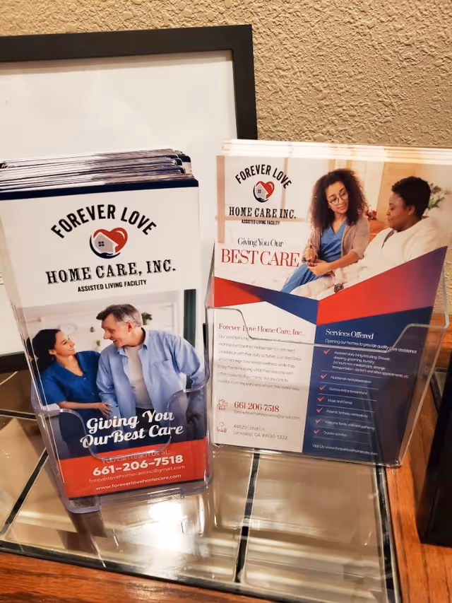 Two brochure holders on a glass table displaying brochures for Forever Love Home Care, Inc., an assisted living facility. The brochures feature images of caregivers interacting with elderly individuals and text promoting their care services.