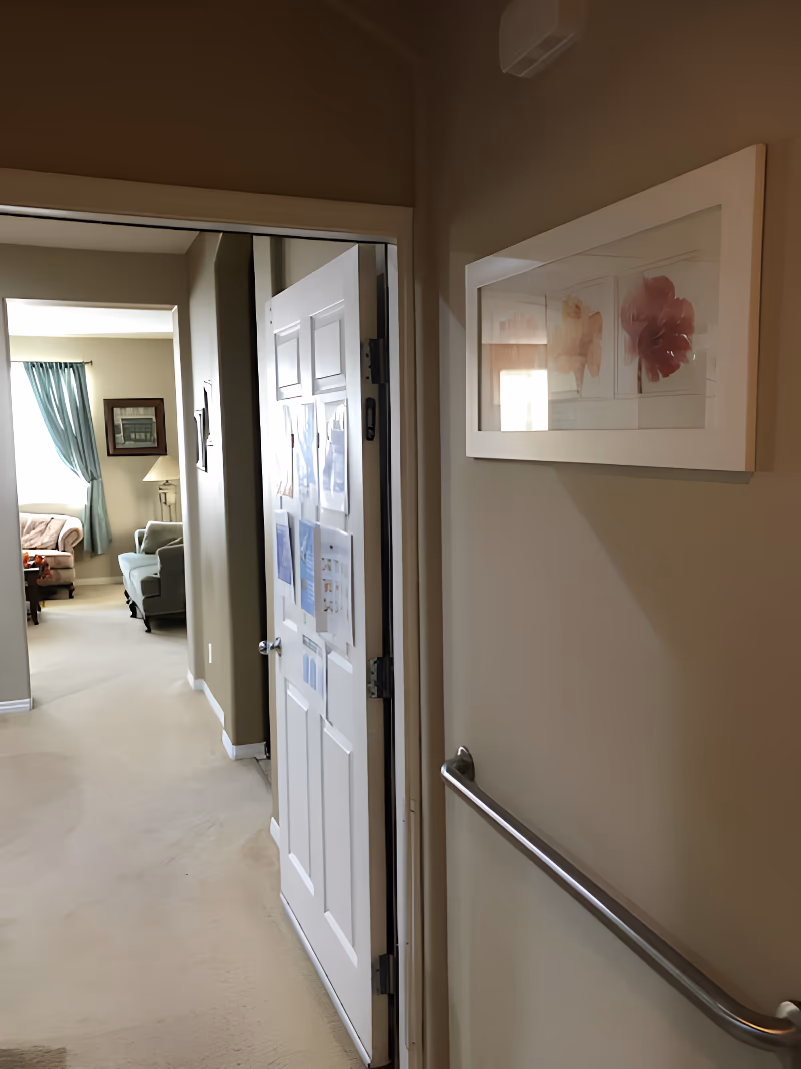 Interior hallway of a senior living unit with a partially open door, a wall handrail, and a view into a sitting area with chairs and curtains.