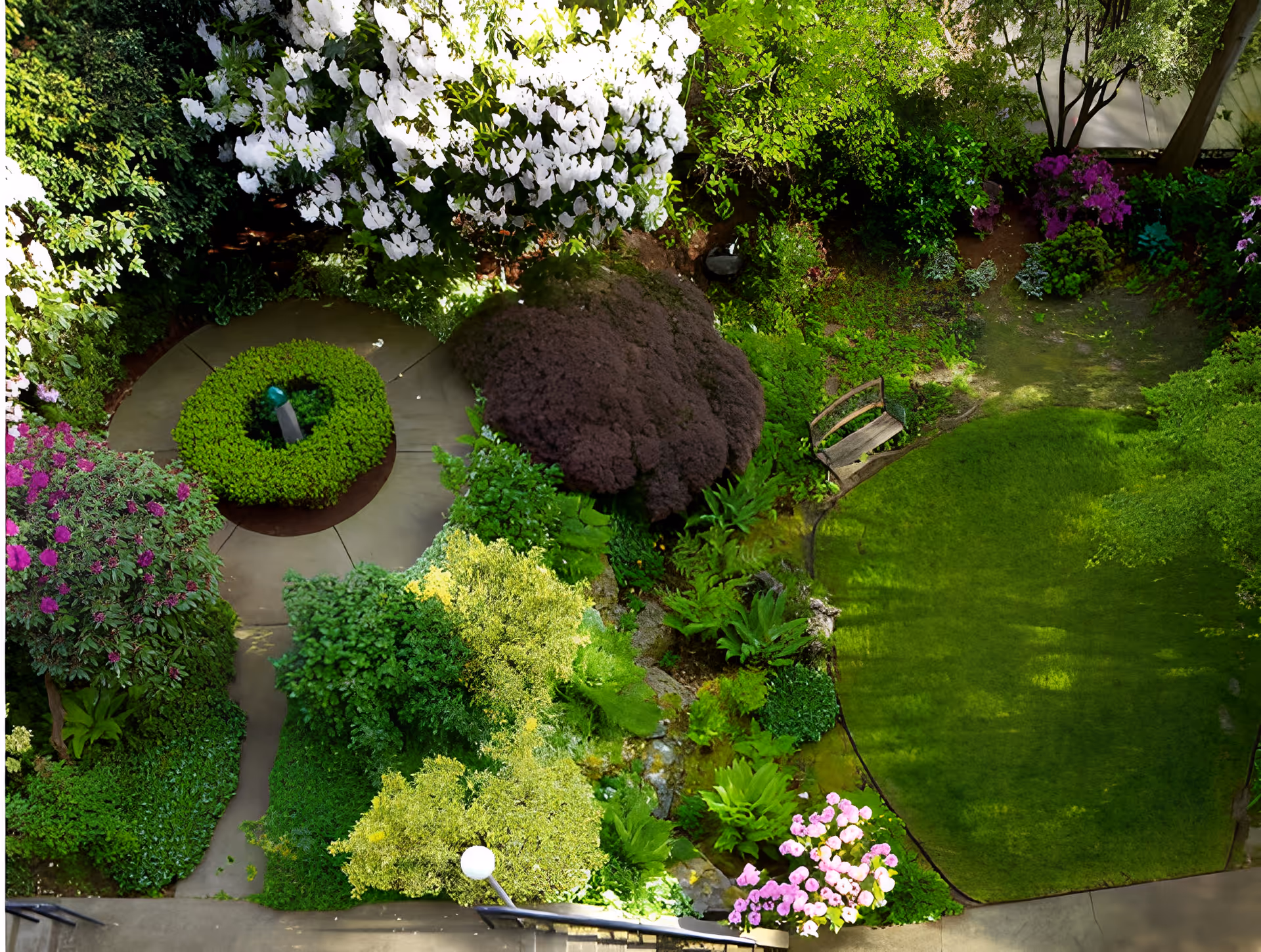 A lush garden area with a variety of green plants, bushes, and colorful flowers. There is a circular paved pathway with a green shrub centerpiece and a small wooden bench on the right side near a grassy lawn. The garden is surrounded by dense foliage and trees.