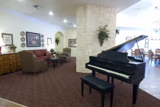 A spacious senior living common room with a black grand piano in the foreground, a stone column, upholstered seating and dining tables in the background.