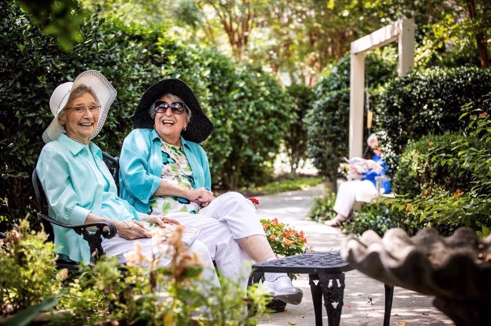 Two elderly women sitting on a bench in a garden area, smiling and wearing sun hats and light clothing. The garden is lush with greenery and flowers, and another elderly person is sitting on a bench further down the path, reading a newspaper.