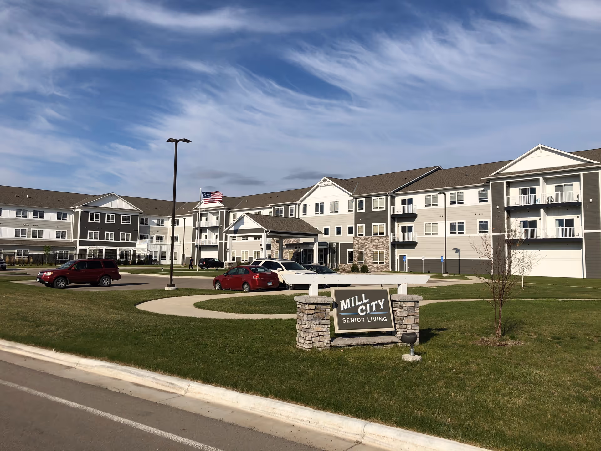 Exterior view of Mill City Senior Living facility showing a large three-story building with balconies, multiple windows, and a covered entrance. There is a green lawn in front with a stone sign displaying the facility's name, several parked cars, a street lamp, and an American flag flying on a flagpole under a partly cloudy sky.