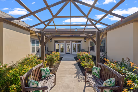 Outdoor courtyard area with a wooden pergola overhead, two wooden benches with green and white patterned cushions facing each other along a concrete pathway, surrounded by bushes and plants, leading to a building entrance with glass doors under a clear blue sky with some clouds.