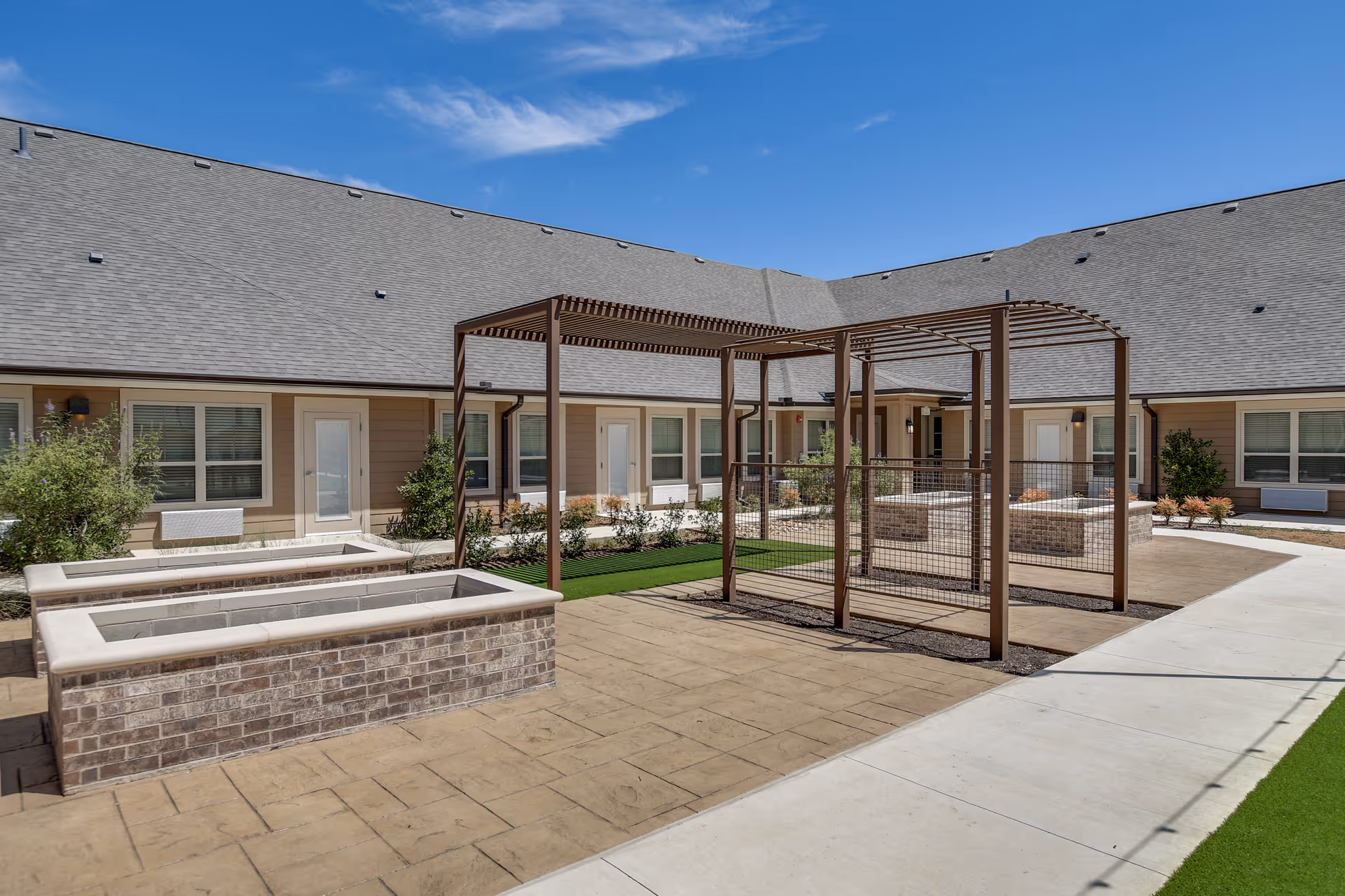 Outdoor courtyard area at a senior living facility with brick raised garden beds, metal pergolas, paved walkways, and beige building walls with multiple windows and doors under a clear blue sky.
