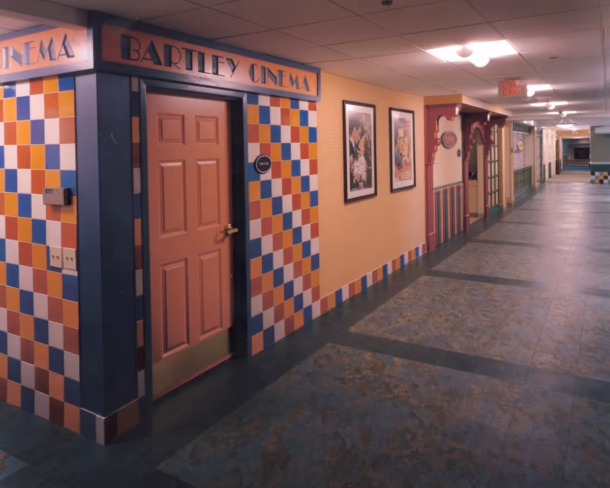 A long indoor hallway with colorful tiled walls and a door labeled 'Bartley Cinema'. The hallway has framed pictures on the wall, decorative wooden trim, and a tiled floor with a pattern. Ceiling lights illuminate the corridor, and an exit sign is visible at the far end.