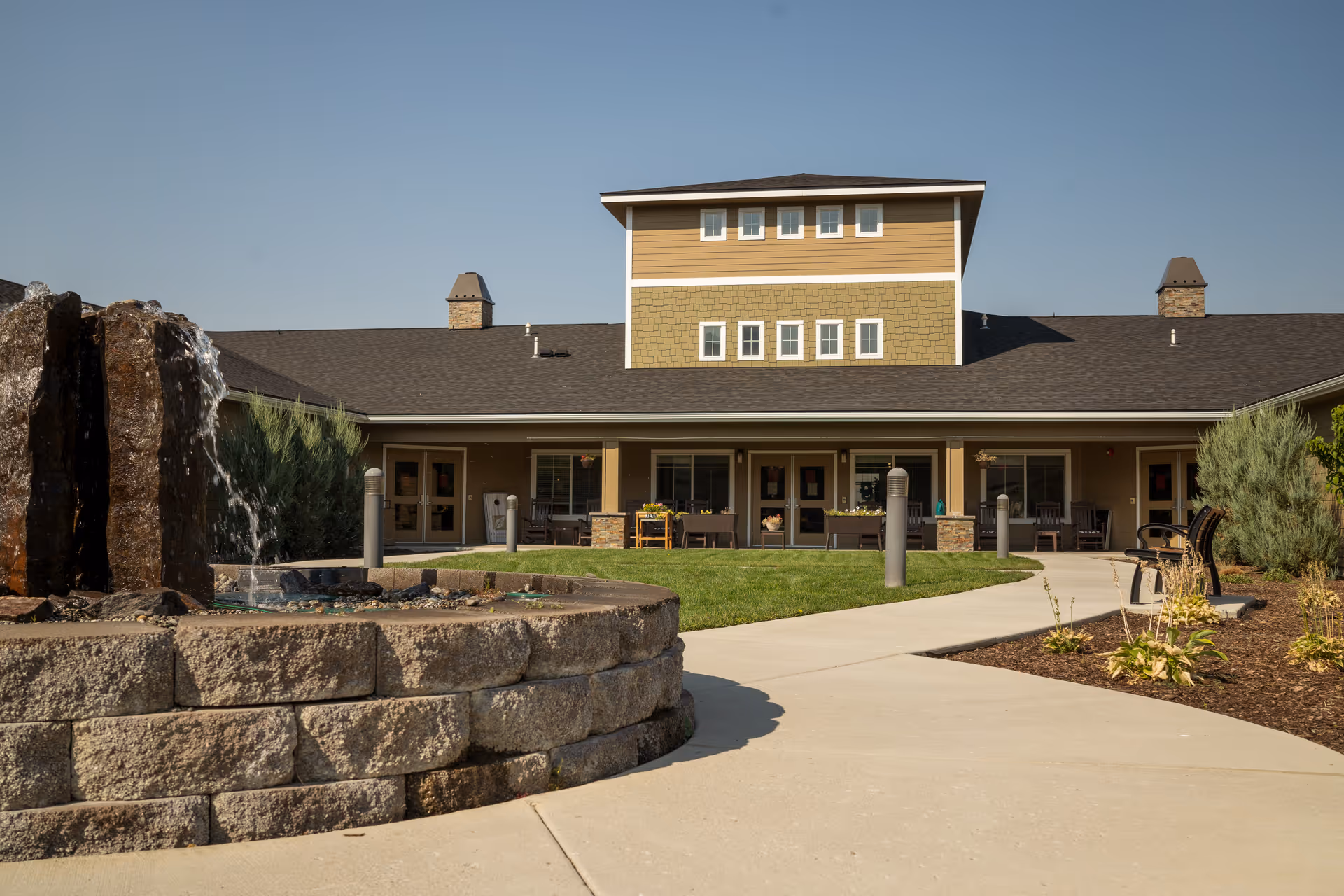 Exterior view of a senior living facility building with a stone water fountain in the foreground, a curved concrete walkway, green lawn, and outdoor seating areas under a covered porch. The building has a two-tone facade with multiple windows and a clear blue sky above.