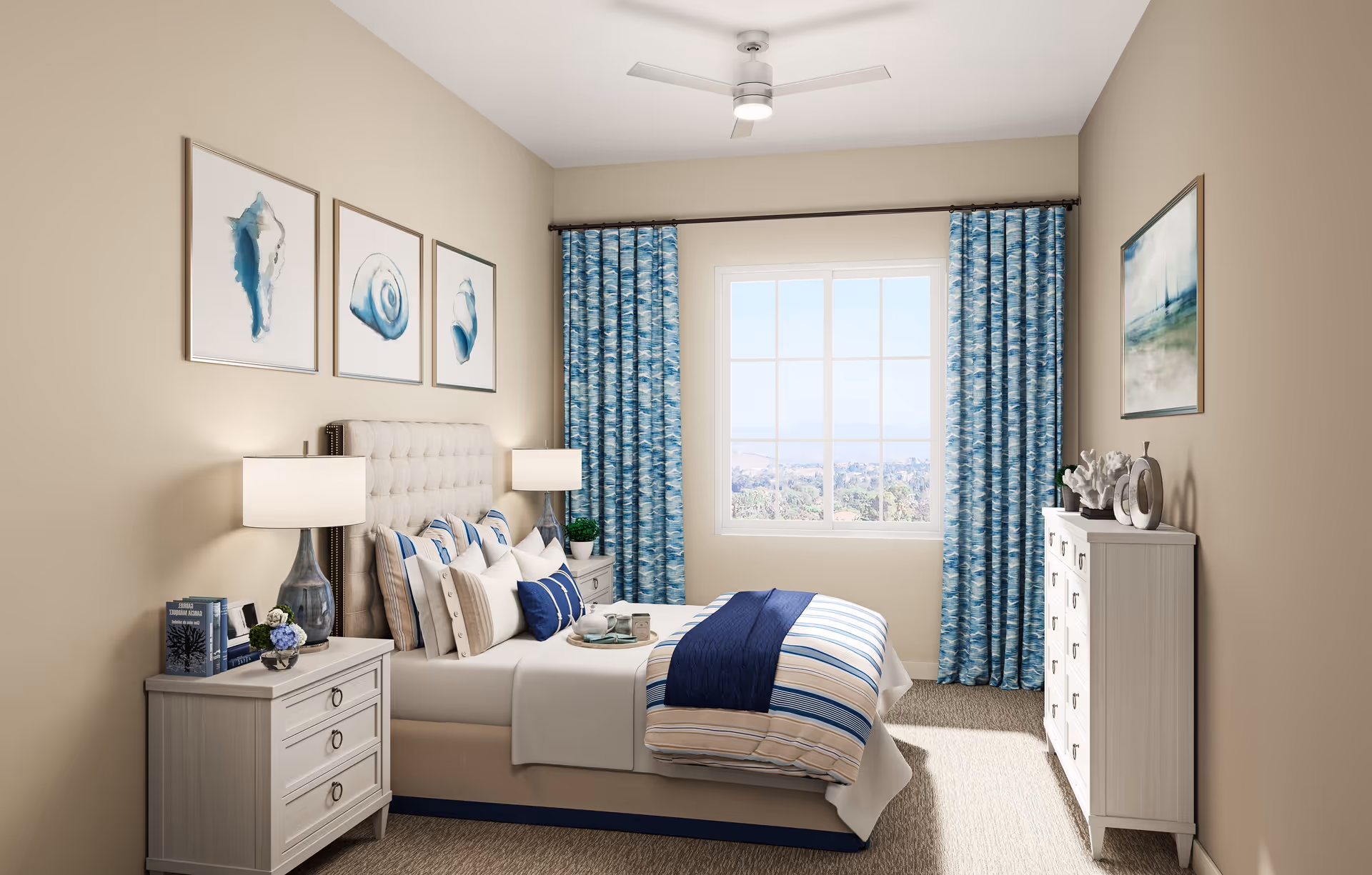 A bright and cozy bedroom with a large window covered by blue patterned curtains. The bed is neatly made with striped bedding in shades of blue, beige, and white, and multiple pillows. There are two bedside tables with lamps and decorative items, and a dresser with art and decor on top. The walls are beige with framed artwork featuring seashells and a landscape painting.