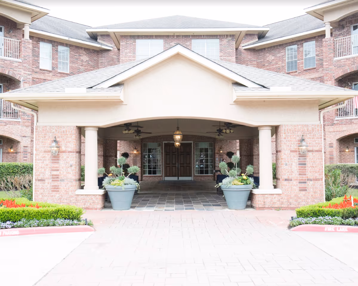 Front entrance of a senior living facility with a covered driveway supported by columns, large potted plants on either side, brick exterior walls, and a double wooden door under a hanging lantern light.
