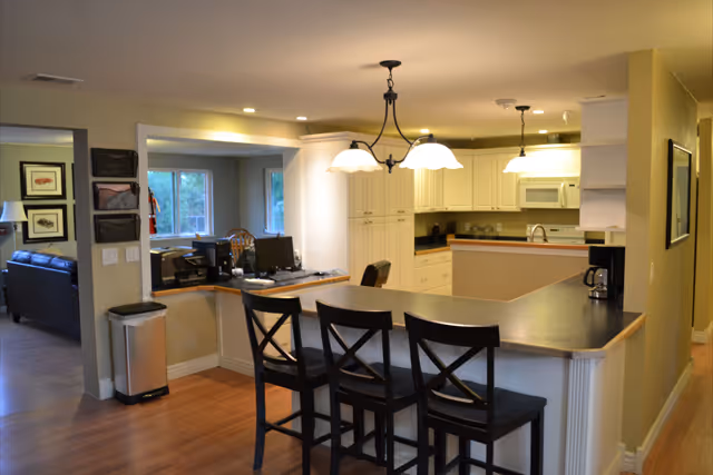 Interior view of a kitchen and dining area in a senior living facility. The kitchen features white cabinets, a microwave, a coffee maker, and a sink. In front of the kitchen counter are three black wooden bar stools. The room is well-lit with ceiling lights and pendant lamps. Adjacent to the kitchen is a small office area with a desk, computer, and chair. The flooring is wooden, and the walls are painted in a light beige color.