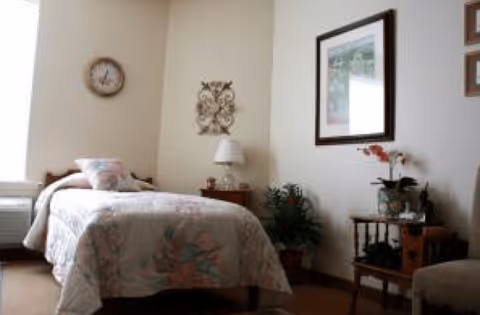 A cozy bedroom with a single bed covered in a floral bedspread, a wooden nightstand with a lamp, a wall clock, decorative wall art, a framed picture, and a small table with plants and flowers.