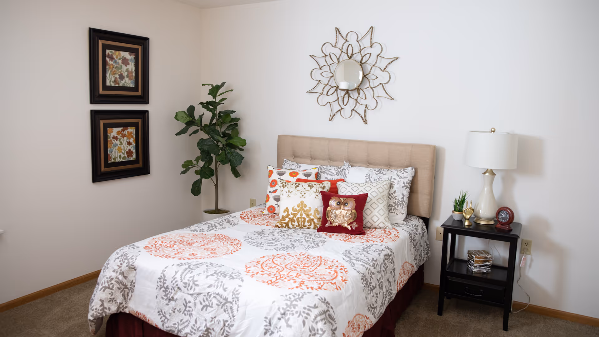 A neatly made bed with a beige upholstered headboard, decorated with multiple patterned pillows including one with an owl design. The bedspread has a white background with orange and gray circular patterns. To the right of the bed is a black nightstand holding a white lamp, a small plant, a clock, and some decorative items. On the wall above the bed is a decorative mirror with a floral metal frame. On the left wall, there are two framed floral artworks stacked vertically. A green potted plant is placed in the corner between the bed and the wall. The room has beige carpet and white walls.