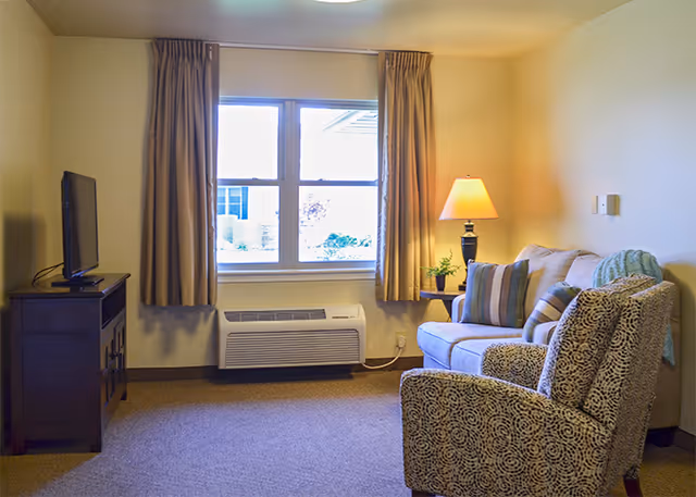 Cozy living room with an armchair and loveseat, a TV on a wooden stand, a window with curtains and a side table lamp.