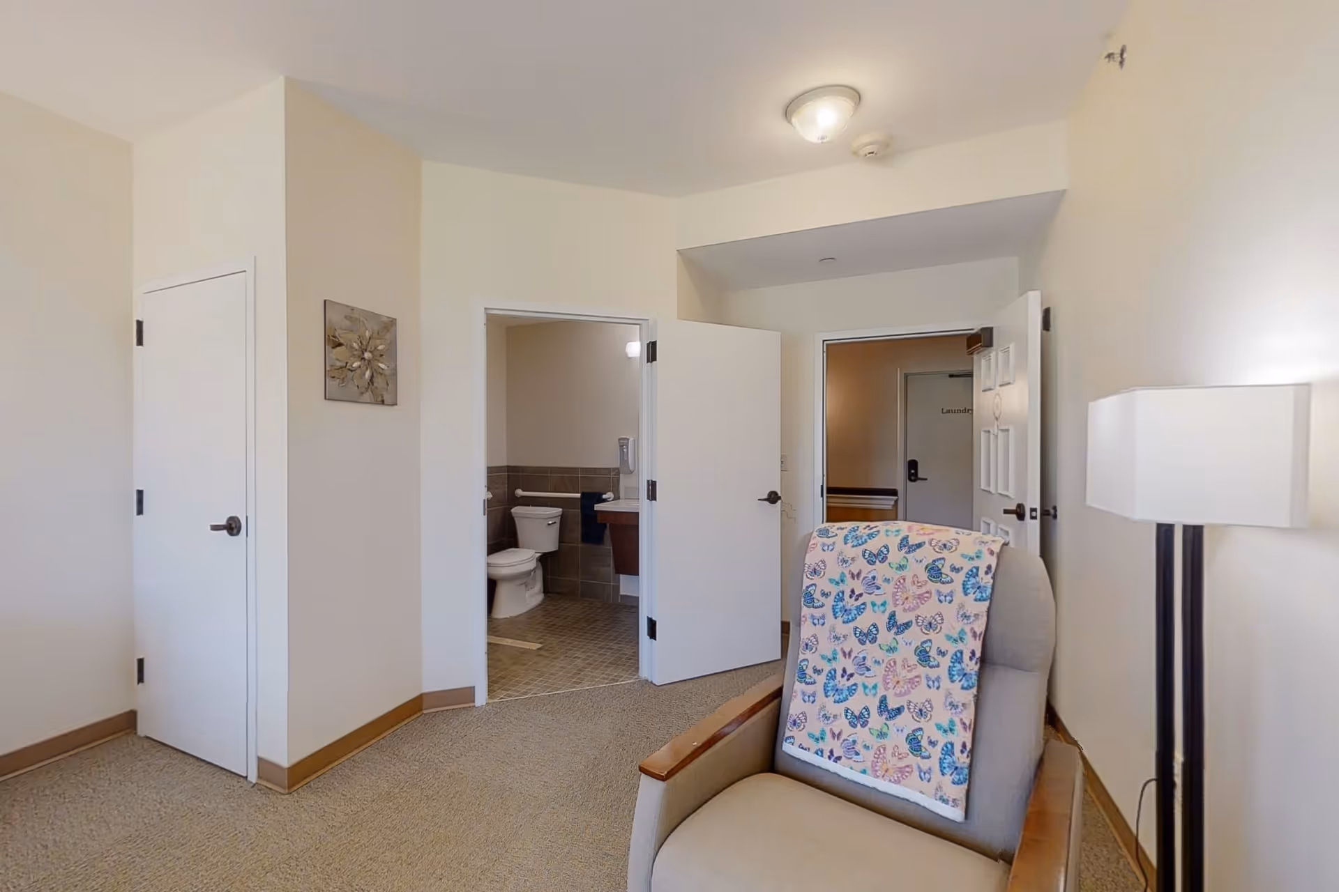Interior view of a senior living facility room showing a beige armchair with a colorful butterfly blanket draped over it, a floor lamp to the right, and two open doors revealing a bathroom with a toilet and sink, and a hallway with a door labeled 'Laundry'. The walls are light-colored and the floor is carpeted.
