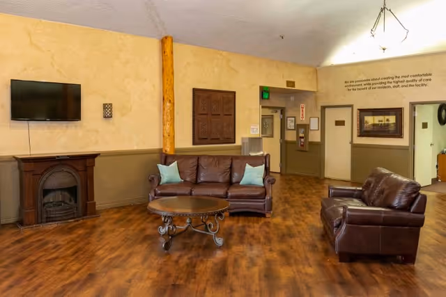 A senior living common room with leather sofas and armchair around a round coffee table, a wall-mounted TV, and wood-look flooring.