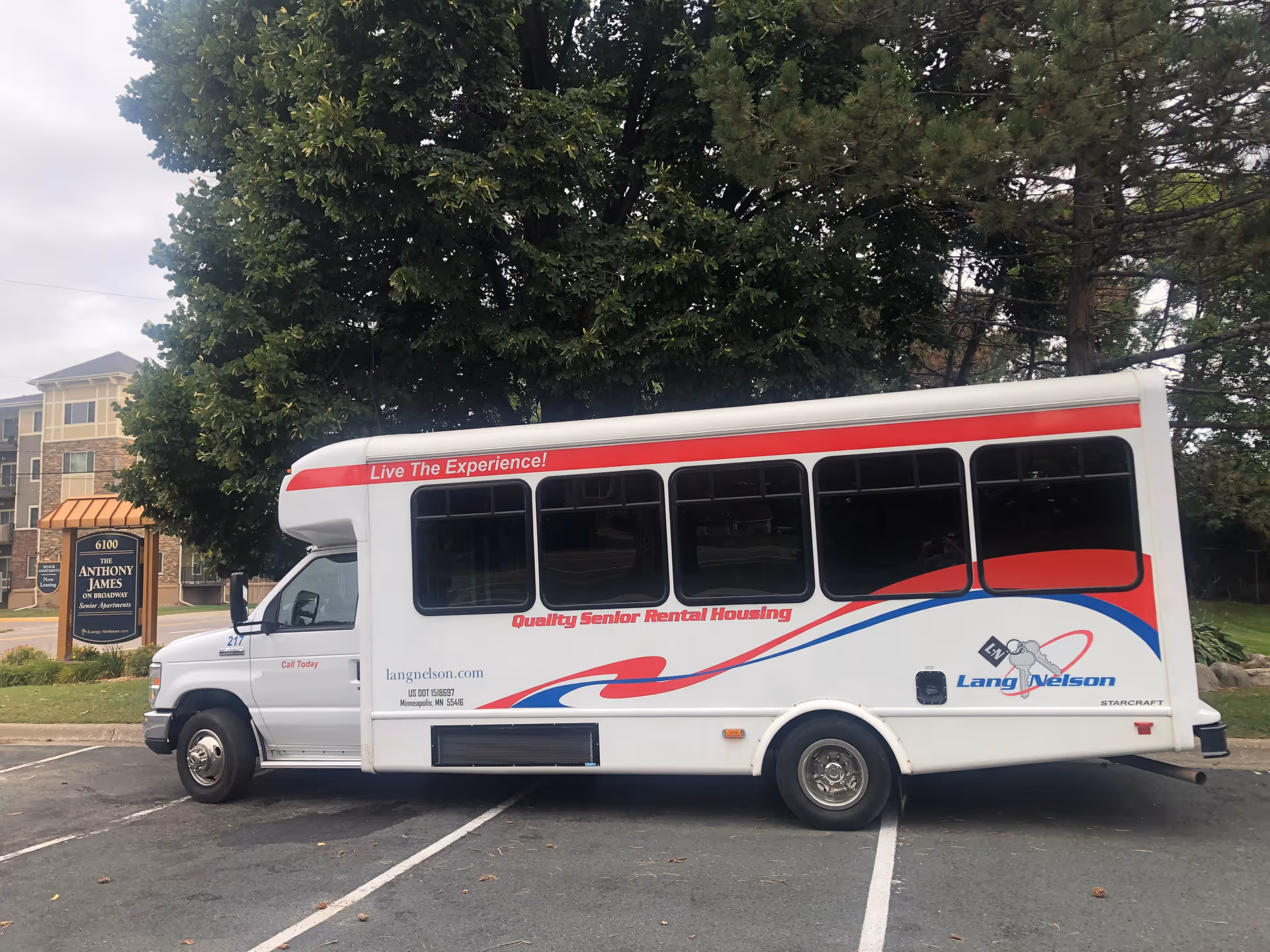 A white shuttle bus with red and blue accents parked in a lot. The bus has text on the side that reads 'Live The Experience! Quality Senior Rental Housing' and 'Lang Nelson'. In the background, there is a sign for The Anthony James senior apartments and a multi-story apartment building partially visible behind trees.