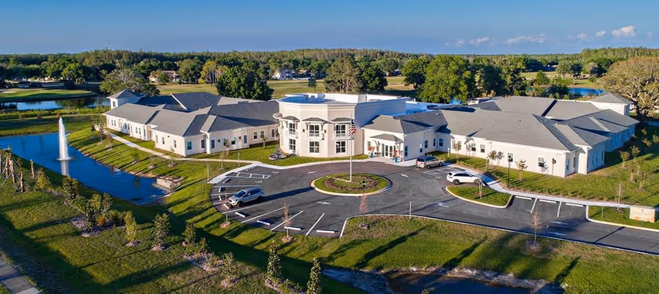 Aerial view of Magnolia Trails at East Lake senior living facility showing a large, single-story building with a circular driveway and an American flag in the center. The building is surrounded by green lawns, trees, a pond with a fountain, and a clear blue sky.
