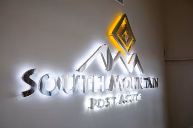 Backlit metallic wall sign reading "South Mountain Post Acute" with a stylized mountain logo mounted on a white interior wall.
