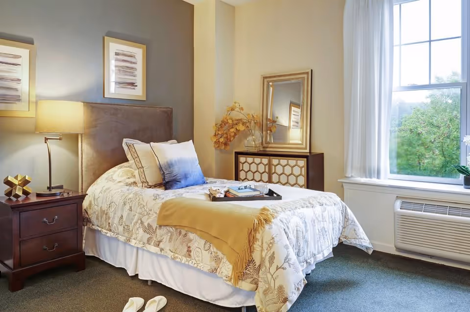 A cozy bedroom with a single bed featuring a floral patterned comforter, multiple pillows, and a yellow throw blanket. Next to the bed is a wooden nightstand with a lamp and decorative item. A dresser with a mirror and a vase with dried flowers is against the wall. A large window with white curtains lets in natural light, and slippers are placed on the carpeted floor near the bed.