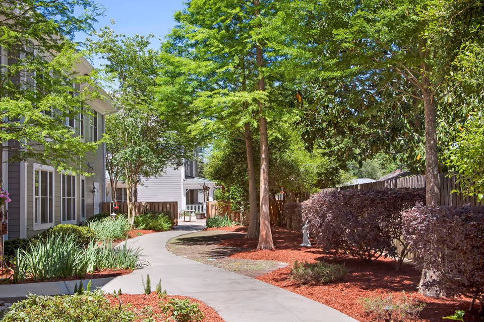 Curved concrete walkway through landscaped gardens and trees between residential buildings at a senior living community.