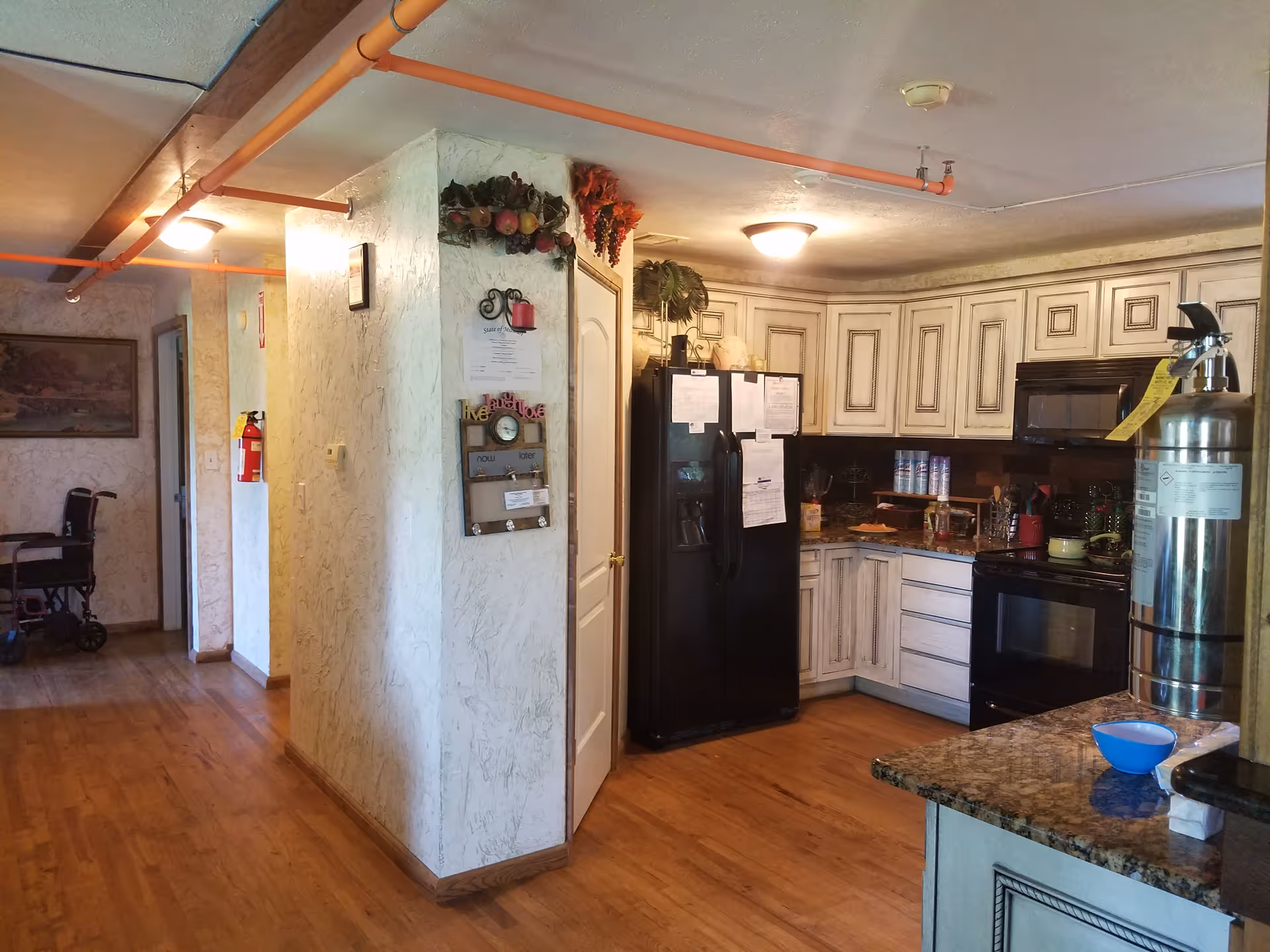 Interior view of a kitchen area in an assisted living facility with wooden floors, white distressed cabinets, a black refrigerator, a black stove, and a granite countertop. There is a fire extinguisher on the right side of the image and a hallway with a wheelchair visible in the background. The walls have a textured finish with decorative items hanging on them.