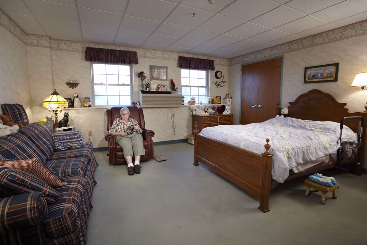 A senior woman sitting in a brown armchair in a cozy bedroom with a wooden bed, a plaid sofa, two windows with valances, and various decorative items including stuffed animals and framed pictures.