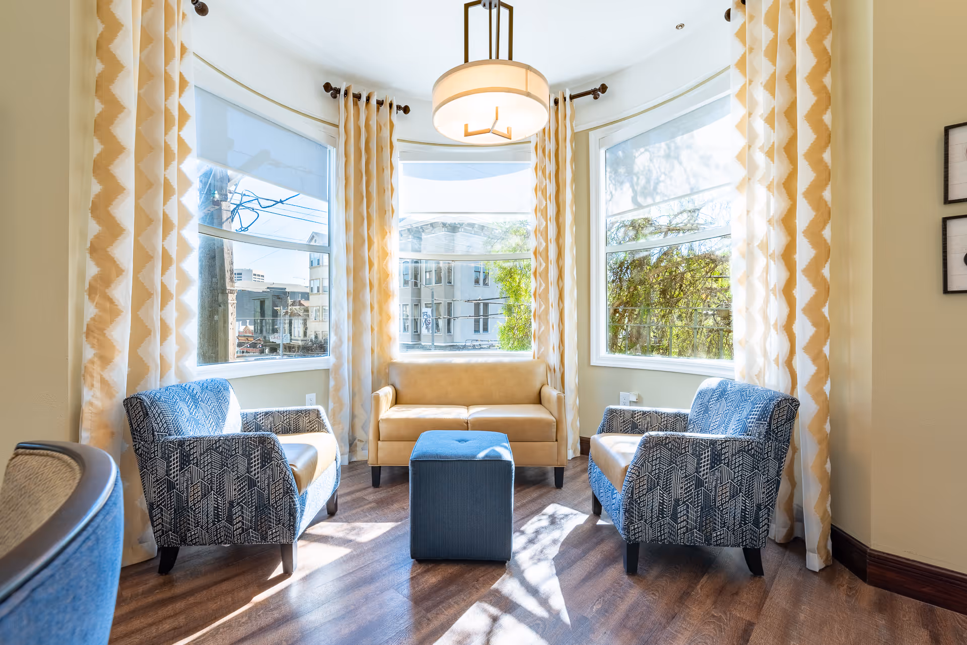Bright seating area with two patterned armchairs, a small sofa and ottoman in front of large bay windows with patterned yellow curtains.