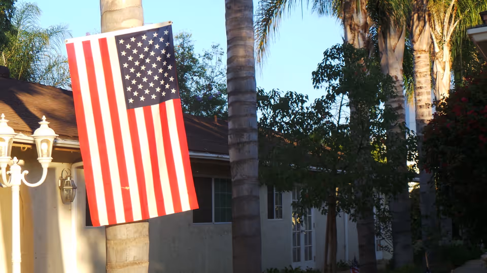 An American flag hanging vertically from a palm tree in front of a single-story building with windows and a door, surrounded by other palm trees and greenery under a clear blue sky.