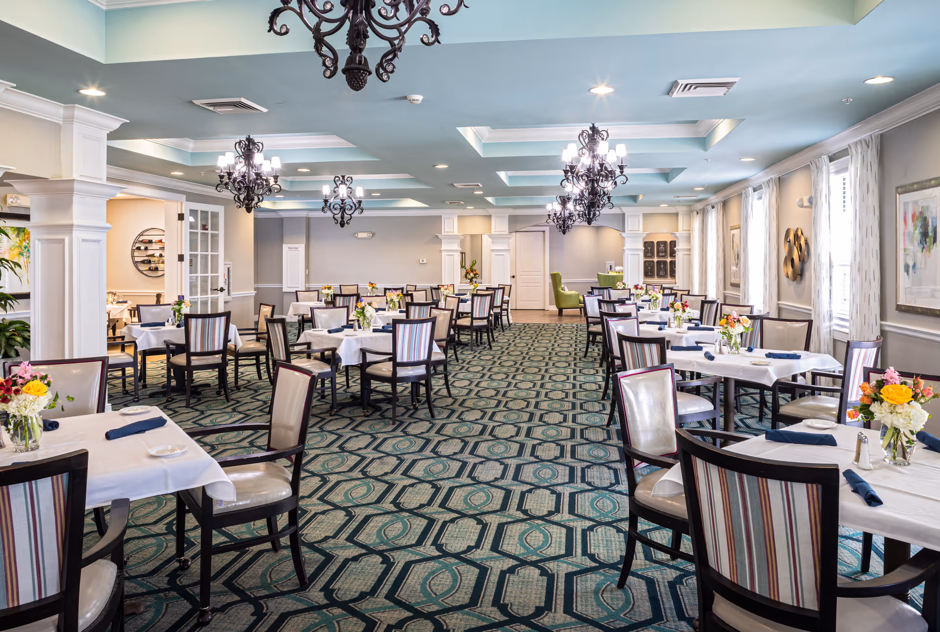 Bright, elegant dining room with multiple tables set with white linens, floral centerpieces, and chandeliers.