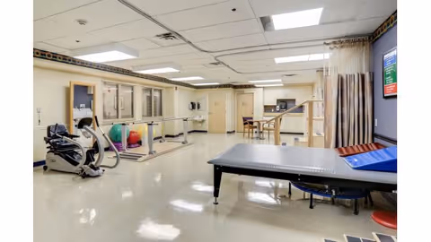 Rehabilitation gym room with exercise equipment, parallel bars, a therapy table, therapy balls, and practice stairs.