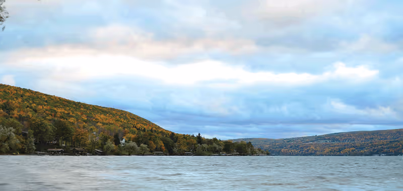A scenic view of a large body of water with forested hills in the background under a cloudy sky.