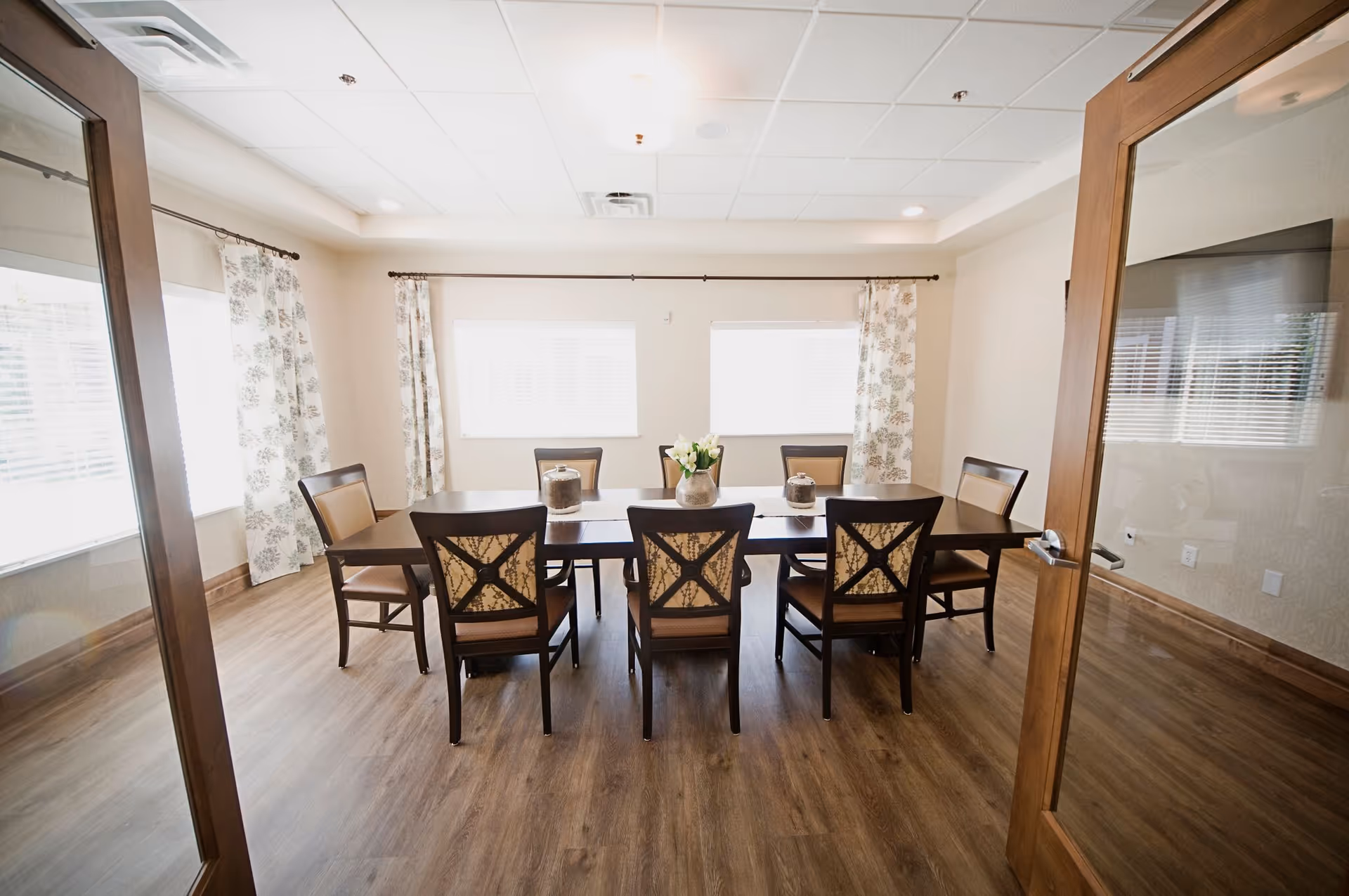 A bright dining room with a rectangular wooden table surrounded by eight chairs. The room has wood flooring, two windows with floral curtains, and a vase with flowers placed in the center of the table.
