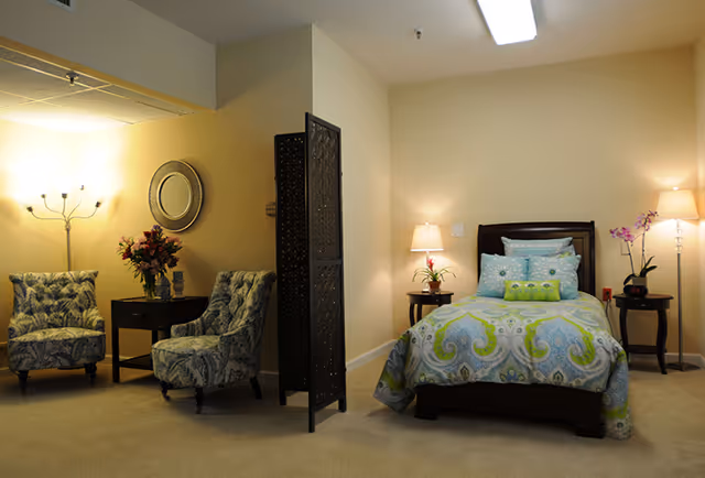 A cozy bedroom with a single bed dressed in blue and green patterned bedding, flanked by two wooden nightstands each with a lamp and a plant. To the left, there are two patterned armchairs with a small table between them holding a vase of flowers, a decorative mirror on the wall above, and a floor lamp providing warm light. A decorative folding screen partially separates the seating area from the bed.