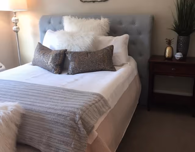 A neatly made bed with a tufted gray headboard, white and fluffy pillows, and two decorative brown pillows. A gray textured blanket is folded at the foot of the bed. To the right of the bed is a wooden nightstand with a small plant, a decorative golden pineapple, and a small round object. A floor lamp with a white shade is visible on the left side of the bed.