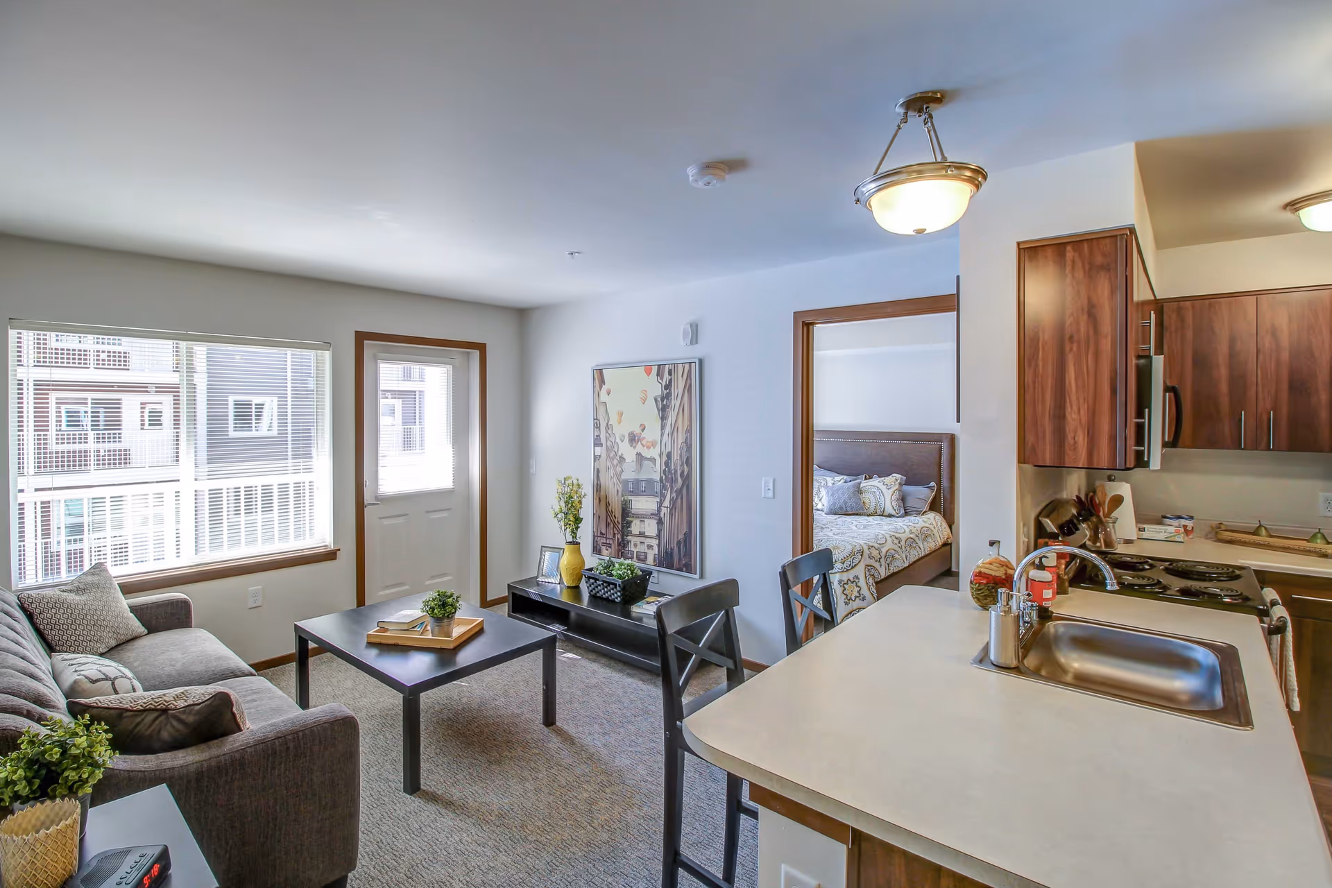 Bright furnished living area with a sofa and coffee table, an open kitchen island with sink, and a bedroom visible through a doorway.