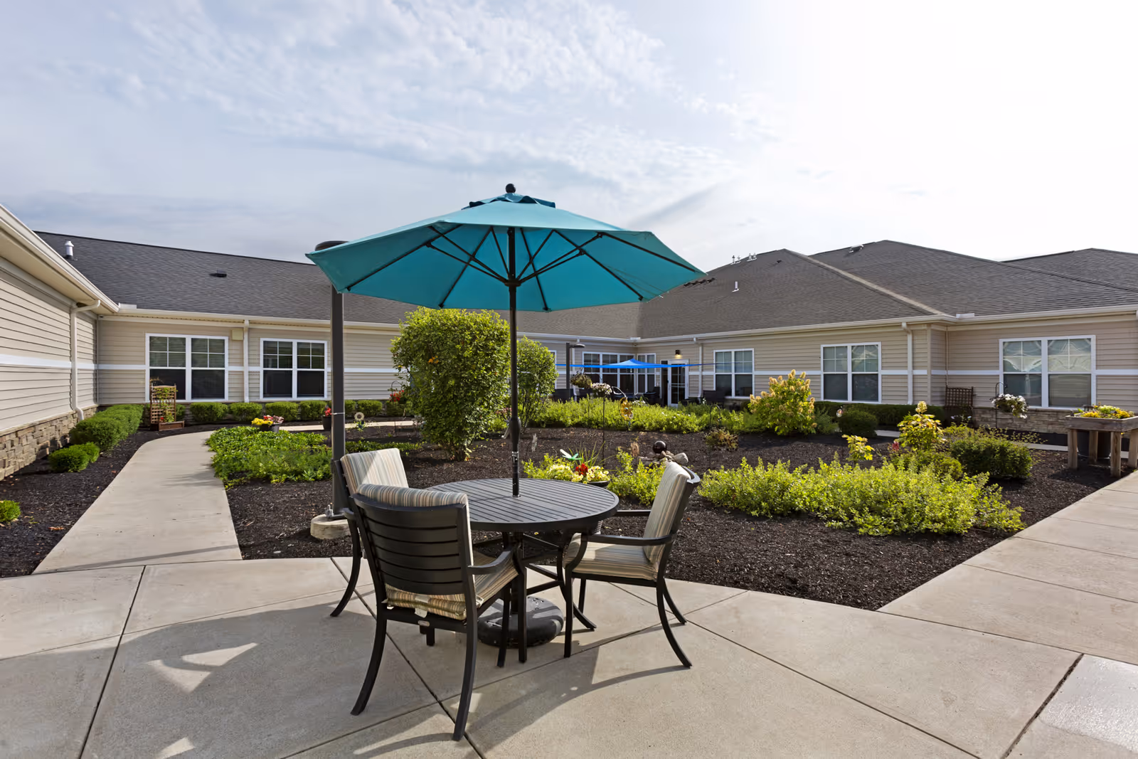 Courtyard patio with a round table, chairs and a teal umbrella in front of a single-story residential building and landscaped garden beds.
