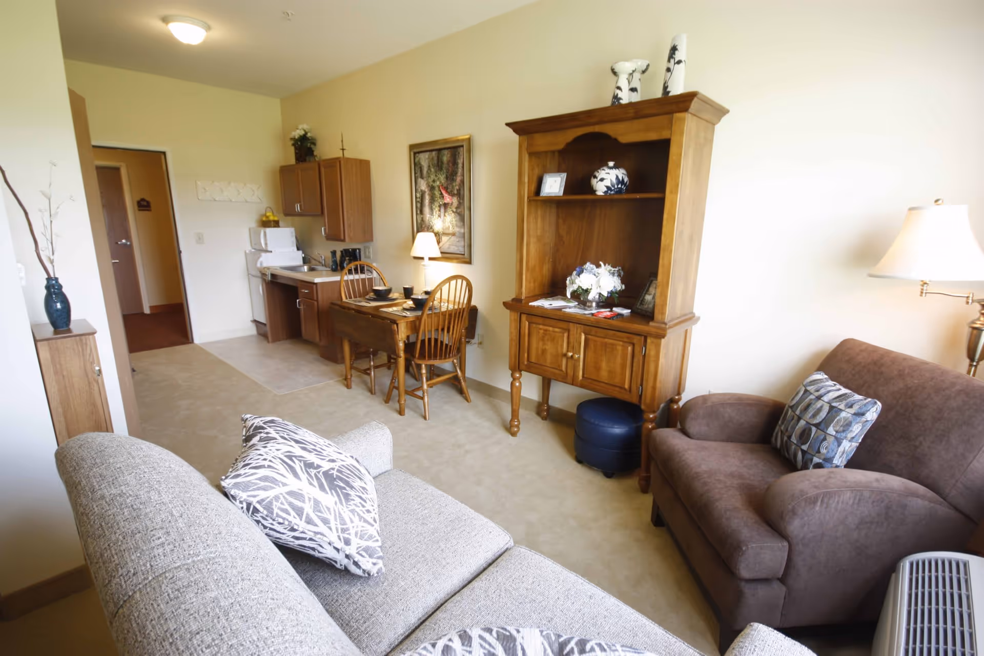 A cozy living area in a senior living facility featuring a gray sofa with patterned pillows, a brown armchair with a decorative pillow, a wooden hutch with decorative items and flowers, a small wooden dining table with two chairs set with dishes, and a kitchenette with a sink, cabinets, and a microwave. The room is softly lit with a table lamp and a ceiling light.