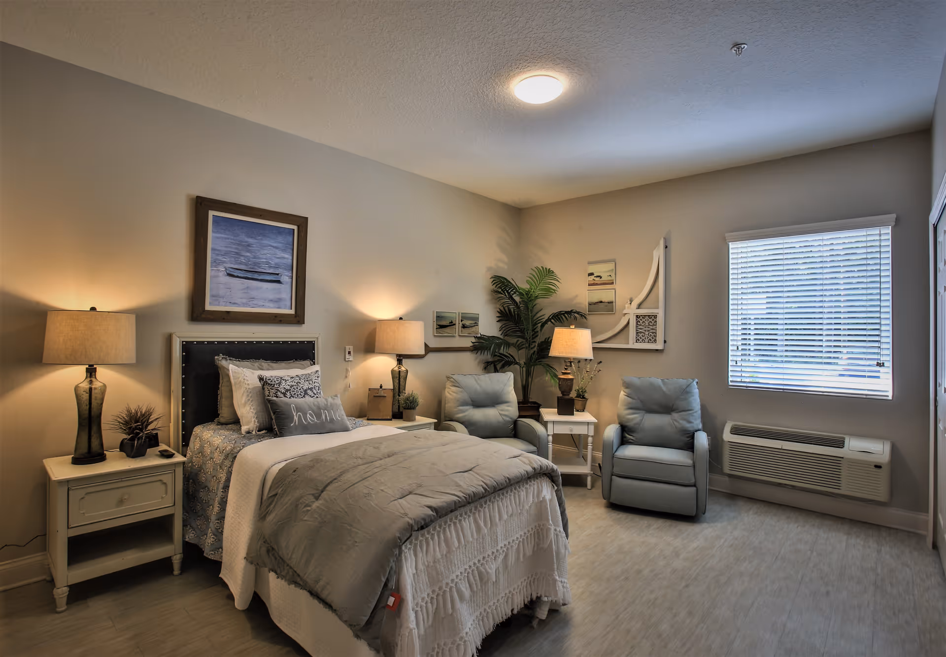 Well-lit bedroom with a made bed, nightstands and lamps, two armchairs, and a window with blinds.