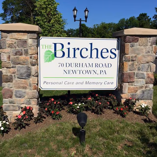 Entrance sign for The Birches at 70 Durham Road, Newtown, PA mounted between two stone pillars with flowers and a lamp post behind it.