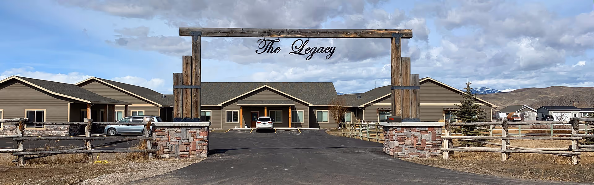 Single-story ranch-style building viewed from the driveway with a wooden entrance arch reading "The Legacy", parked cars, split-rail fencing and distant mountains.