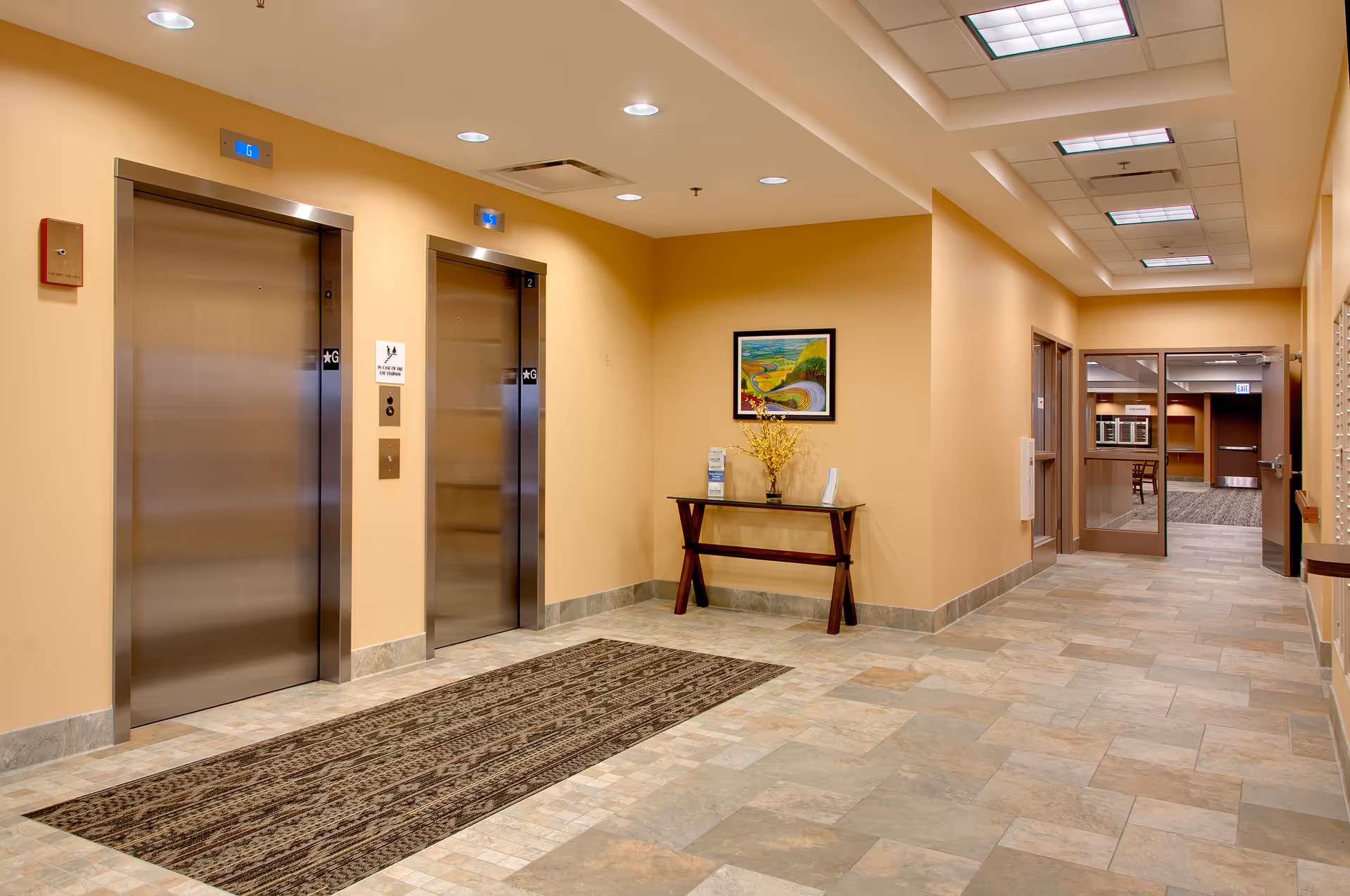Interior hallway of a senior living facility with two closed stainless steel elevators on the left, a patterned rug in front of them, beige walls, tiled floor, a small table with a vase of yellow flowers and brochures, and a framed landscape painting above the table. The hallway extends to the right with glass doors leading to another room.