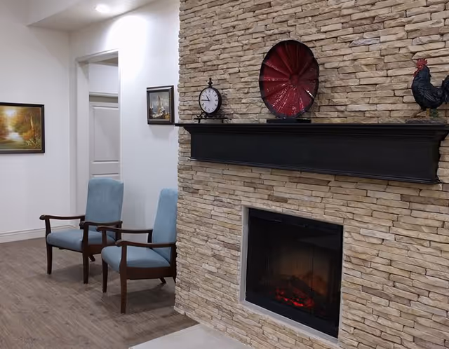 A cozy interior corner featuring a stone fireplace with a black mantel displaying a clock, a red decorative plate, and a rooster figurine. Two light blue upholstered wooden armchairs are positioned against a white wall with two framed paintings. The floor is wood with a light-colored rug partially visible.