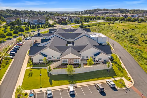 Aerial view of Poet's Walk Leesburg, A Memory Care Community, showing a large, single-story building with multiple roof sections, surrounded by green lawns, parking spaces with several cars, and nearby residential areas under a clear blue sky.